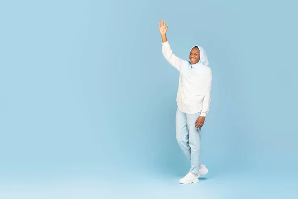 Full body shot of black muslim woman wearing hijab, walking and waving hand isolated on blue background, free space. Lady greeting with someone