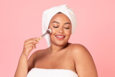 Makyaj fırçasıyla havluya sarılmış çekici, genç, tombul bir kadın yanaklarına allık sürüyor, pembe stüdyo arka planında yepyeni makyaj ürünlerinin tadını çıkarıyor.