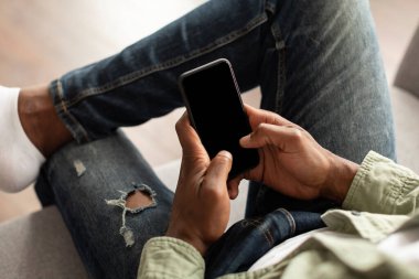 Olgun bir Afro-Amerikan erkeği kanepeye oturup akıllı telefondan oda içi boş ekranlı video izler. Yeni sohbet uygulaması, çalışma, öğrenim ve iletişim için aygıt, web sitesi, iyi bir internet teklifi ve evde reklam