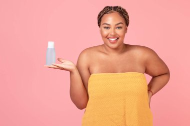 Vücut pozitifliği olan Afro-Amerikan kadın elinde su şişesi, yeni kozmetik ürünlerin reklamını yapıyor, pembe arka planda poz veriyor, stüdyoda çekim yapıyor.
