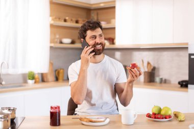 Beyaz tişörtlü, sakallı yetişkin bir beyaz adamın sandviç yemesine ve modern mutfağın içinde telefonla konuşmasına sevindim. Günaydın ve kahvaltı, iletişim, haber ve dedikodu, harika bir teklif ve evde reklam