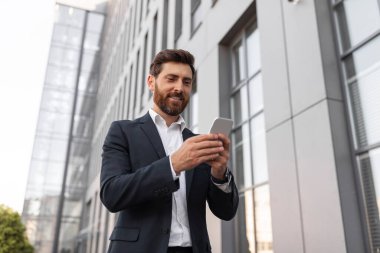 Takım elbiseli mutlu, kendine güvenen genç Avrupalı patron daktilo ediyor, şehir caddesindeki binanın yakınında telefonla konuşuyor. Gadget, iş, finans ve girişim uygulaması, modern iş, kariyer, reklam ve teklif