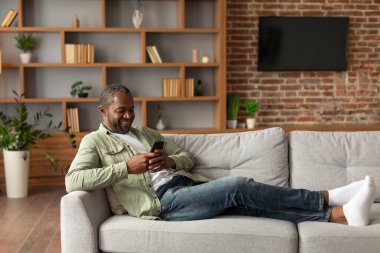 Gözlüklü, gülümseyen Afro-Amerikan erkeği akıllı telefondan mesaj okuyor, sohbet ediyor, oturma odasındaki kanepede video izliyor. Sosyal ağlar, blog, internette arama ve evdeki aygıtlar için uygulama