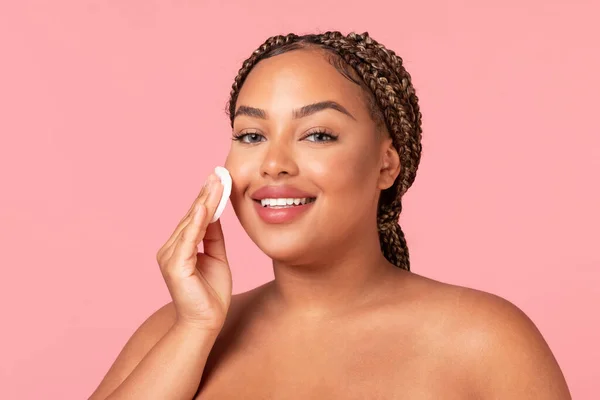 Facial skincare concept. Happy black chubby woman using cotton pad, caring for smooth skin, looking and smiling at camera, posing over pink studio background. Beauty and cosmetics concept