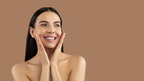 Aesthetic medicine and cosmetology concept. Happy beautiful brunette young woman touching her perfect face skin, posing naked over beige studio background, looking at copy space and smiling, panorma