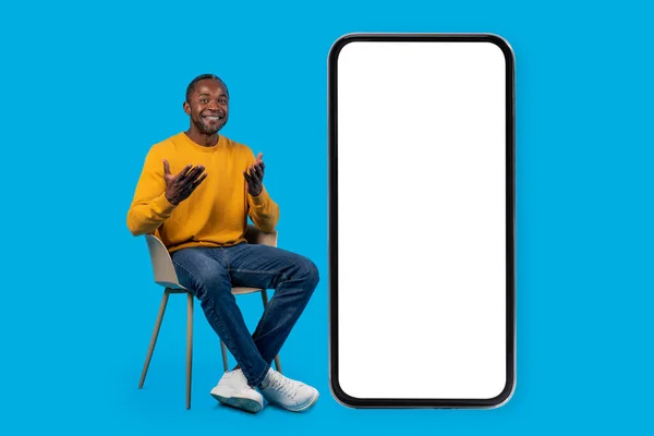 快乐而英俊的中年黑人男子 坐在长椅上 坐在白色空屏幕的大手机旁 笑容满面 做手势 网上拍卖的模型 蓝色工作室背景 — 图库照片