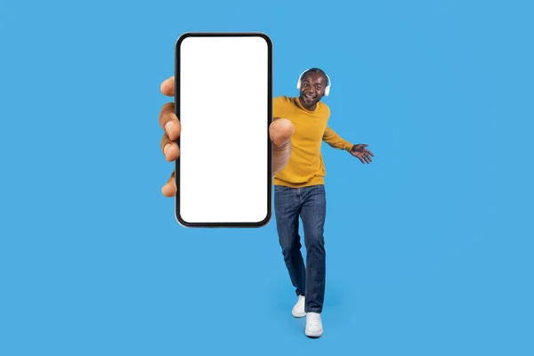 快乐成熟的非洲裔美国男人 身穿时髦休闲装 听音乐和跳舞 使用现代无线耳机 展示带有模拟屏幕的智能手机 蓝色工作室背景 复制空间 — 图库照片