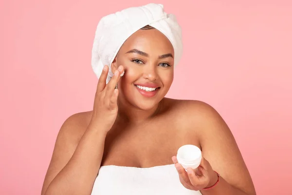 美しいアフリカ系アメリカ人の体の正の女性のクリームの瓶を保持し ピンクの背景にポーズをとり 頬に保湿剤を適用します 毎日の治療 自宅での美容ケアのための化粧品 — ストック写真