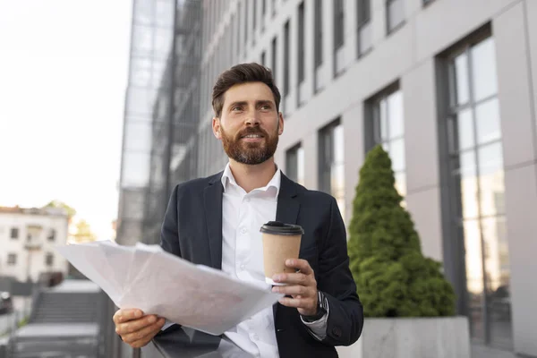 Cheerful confident young european guy boss with beard in suit with documents, enjoy cup of takeaway drink looks up at empty space on balcony of building. Coffee break contract, business, ad and offer