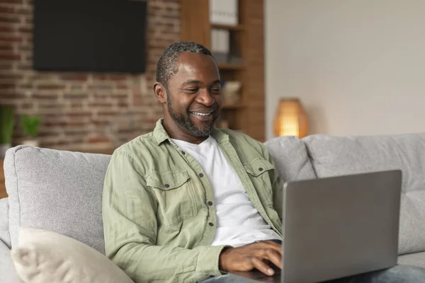 嬉しい陽気な中年アフリカ系アメリカ人男性のソファの上に座ってノートパソコンでビデオを見て リビングルームのインテリアでオンラインコールしています インターネットでサーフィンしたり ブログをしたり 自宅でソーシャルネットワークでチャットしたり — ストック写真