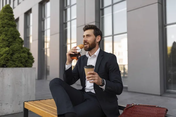Happy hangry young european male manager with beard in suit with cup of coffee takeaway eats croissant, sits on bench near building in city. Food, lunch outdoor, break from work and rest at free time