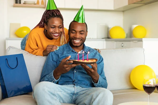 Happy african american wife surprising her husband with handmade birthday pie, man blowing candles and smiling, enjoying b-day celebration at home, free space