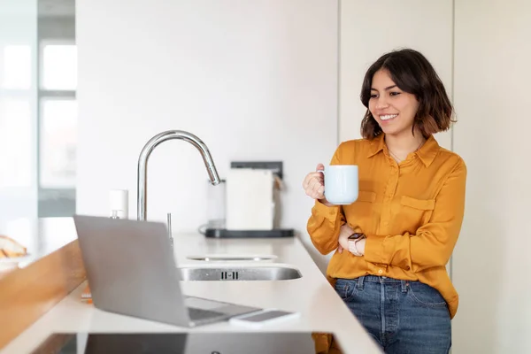 Young Smiling Arab Woman Using Laptop And Drinking Coffee In Kitchen, Happy Millennial Middle Eastern Female Watching Videos On Computer And Enjoying Caffeine Drink At Home, Copy Space
