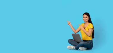 Bin yıllık Asyalı blogcu kız öğrencinin yerde oturup bilgisayarın boş yeri göstermesine sevindim. Mavi arka planda, panoramada, stüdyoda izole edilmiş. Çalışma ve öğrenim için malzeme, satış, reklam ve teklif