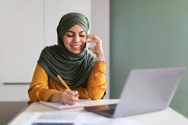 携帯電話で話すヒジャブ語のムスリム女性を笑顔にし 情報をメモし 携帯電話で話す若いイスラム教徒女性のフリーランサーと自宅のオフィスでデスクに座っている間にメモを取ります — ストック写真