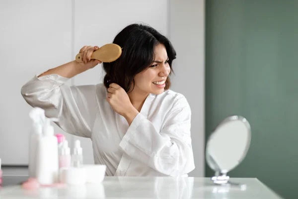 stock image Hair detangling concept. Angry middle eastern young lady in silk robe brushing dry hair with wooden hairbrush, suffer from pain during beauty routine, sitting at beauty table at home, look at mirror