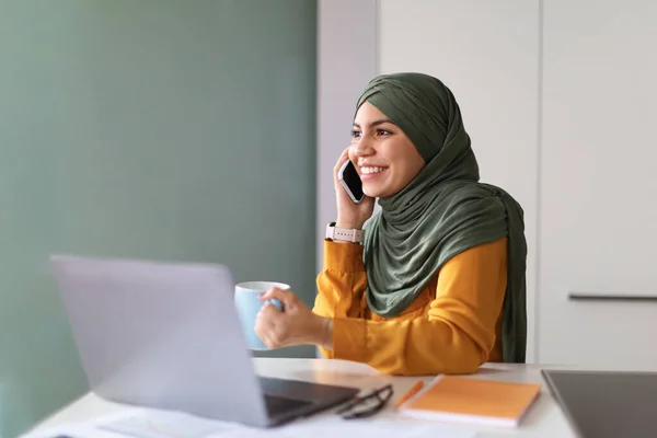 職場での携帯電話で話すムスリム女性の笑顔とコーヒーを飲む職場では 若いイスラム教徒女性がノートパソコンを持ってデスクに座ってモバイル会話をし 仕事で休憩を楽しみます — ストック写真