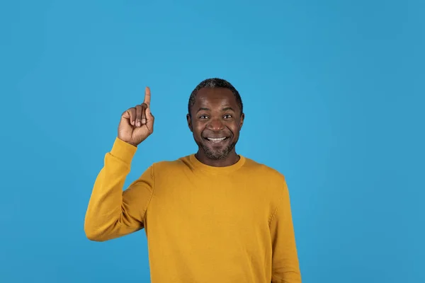 黄色のセーターを着た幸せな中年のアフリカ人男性は アイデアやEurekaジェスチャーや笑顔を示す上を指して 興味深い計画を生成します スタジオは青の背景を撮影し スペースをコピー — ストック写真