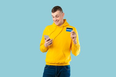 Akıllı telefon ve kredi kartıyla online alışveriş yapan mutlu Avrupalı adam. İnternetten ödeme yapıyor. Mavi stüdyo arka planında duruyor. E-ticaret kavramı