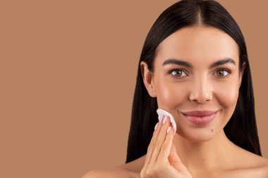 Güzel, esmer, uzun saçlı, pamuk pedi kullanan ve gülümseyen, bej renkli stüdyo arka planında izole edilmiş çıplak bir kadının yakın plan portresi. Yüz temizleme, cilt bakım konsepti