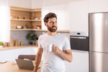 Beyaz tişörtlü, sakallı, düşünceli, düşünceli, olgun, beyaz bir beyaz adam, fincanda içmekten hoşlanıyor, mutfak içinde boş bir alana bakıyor. Başlangıç, iş ve iş için evde fikir, büyük reklam ve teklif