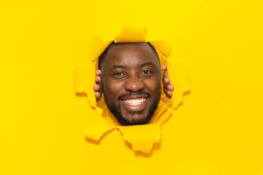 Mutlu Afro-Amerikalı adam sarı kağıt üzerinde poz veriyor. Stüdyo arka planındaki delikten kameraya bakıp gülümsüyor. Harika bir reklam konsepti. Kolaj