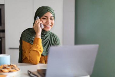 Evde dizüstü bilgisayar kullanan ve cep telefonuyla konuşan gülümseyen çok yönlü kadın, başörtüsü takmış bilgisayar üzerinde çalışan ve mutfaktaki masada otururken cep telefonu konuşması yapan genç İslami kadın.