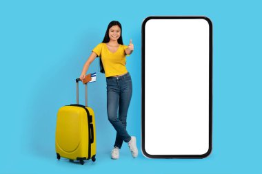 Sarı tişörtlü, bavullu neşeli genç Japon bayan. Biletler, mavi arka planda izole edilmiş, boş ekranlı, dev bir telefonun yanında baş parmak gösteriyor. Seyahat, tatil, gezi, reklam ve teklif uygulaması