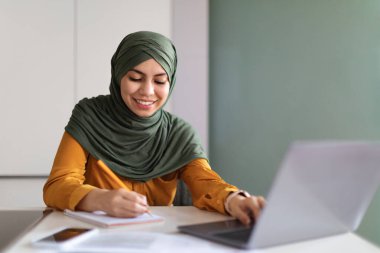 Online Eğitim Konsepti. Genç tesettürlü Müslüman kadın evde dizüstü bilgisayarla tesettüre giriyor, İslami bayan bilgisayar kullanıyor ve not alıyor, uzak öğrenimin ve boş alanın tadını çıkarıyor.