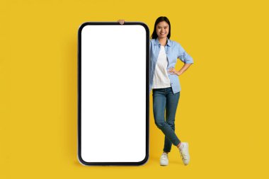 Neşeli, bin yıllık Koreli bayan öğrenci, boş ekranlı, sarı arka planda, stüdyoda izole edilmiş büyük bir akıllı telefonuyla gündelik ayakta duruyor. Satılık aygıt, serbest çalışma ve öğrenim uygulaması, reklam ve teklif