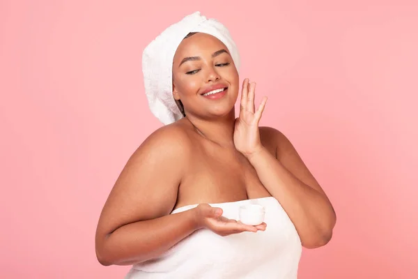 スキンケアの概念 幸せな黒オーバーサイズ女性でタオルで頭の上に適用保湿顔クリーム 美しいアフリカ系アメリカ人肥満女性立ってピンクの背景 — ストック写真