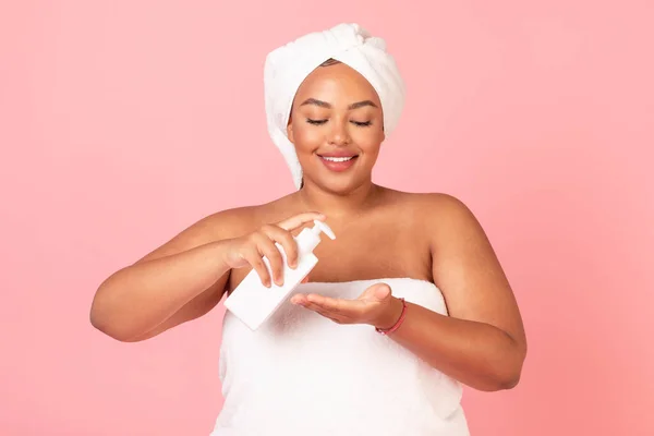 体の栄養の概念 幸せな黒太りすぎの女性を適用保湿化粧水オン手 ポーズタオルに包まれたピンクのスタジオの背景 — ストック写真