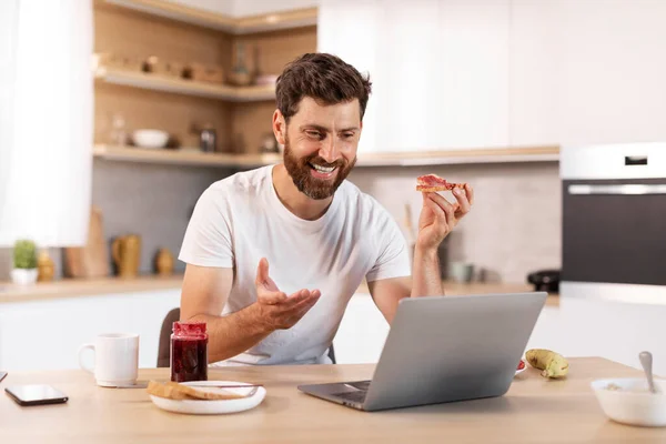 面带微笑的中年白人男性 留着胡子 穿着白色T恤 吃三明治 有视频通话 在厨房内部摆姿势 早上好 远程见面 在家里的社交距离 — 图库照片