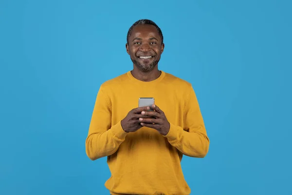 黄色の保持スマートフォンで幸せなハンサムなアフリカ系アメリカ人の成熟した男とカメラで笑顔 最新のモバイルアプリを楽しんで 女性とオンラインでチャット 青のスタジオの背景に隔離された コピースペース — ストック写真