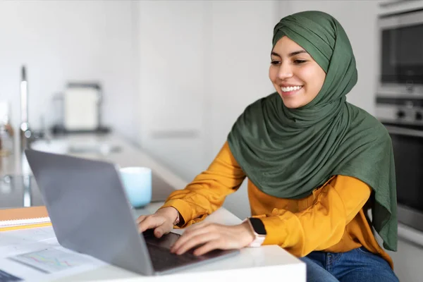 リモートワーク 自宅で働いている間 ラップトップを使用してヒジャブで幸せな若いイスラム教徒の女性のフリーランサー キッチンでデスクで座っているイスラム教徒の女性を笑顔とコンピュータキーボードを入力し フリースペース — ストック写真