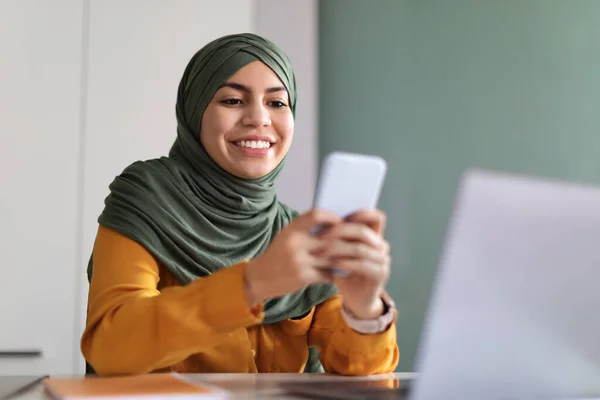 在智能手机上浏览应用程序的阿拉伯女性坐在办公桌前 年轻的穆斯林女性在网上购物或交流时使用手机 在有空余空间的情况下拍照 — 图库照片