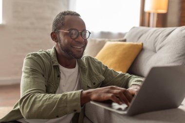 Görüntülü arama ve iş. Neşeli orta yaşlı gözlüklü Afro-Amerikan erkek web kamerası bilgisayarına bakıyor, oturma odasının içindeki sosyal ağlarda yazışıyor ve sohbet ediyor. Uzaktan iş, evde yeni normal.