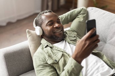 Orta yaşlı, kablosuz kulaklıklı Afro-Amerikan erkek müzik dinliyor, akıllı telefondan yazı yazıyor, oturma odasında boş vakitlerin tadını çıkarıyor. Ses uygulaması ile dinlenme ve dinlenme, evde video izleme