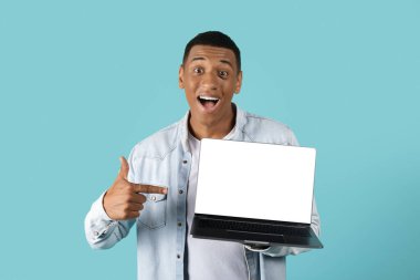 Ağzı açık, boş ekranlı, mavi arka planda izole bir şekilde dizüstü bilgisayarı işaret eden heyecanlı, Afro-Amerikalı genç adam şok oldu. Çalışma, iş ve iş kumandası, online zafer, teklif ve reklam