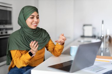 Telekonferans Konsepti. Gülümseyen müslüman kadın evde dizüstü bilgisayarla video görüşmesi yapıyor, tesettürlü Arap kadın konuşuyor ve kameraya işaret ediyor, çevrimiçi iletişimden keyif alıyor