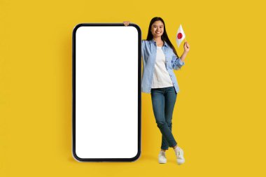 Japonya 'nın neşeli Asyalı kız öğrenci bayrağı boş ekranlı, sarı arka planda, stüdyoda izole edilmiş büyük akıllı telefonlu. Seyahat, dil öğrenimi, değişim eğitimi için uygulama ve aygıt