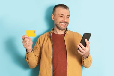 Mobil alışveriş konsepti. Akıllı telefon ve kredi kartı kullanan mutlu olgun bir adam mavi stüdyo geçmişi üzerinden ödeme yapıyor. Bankacılık uygulaması ve ekommerce kavramı