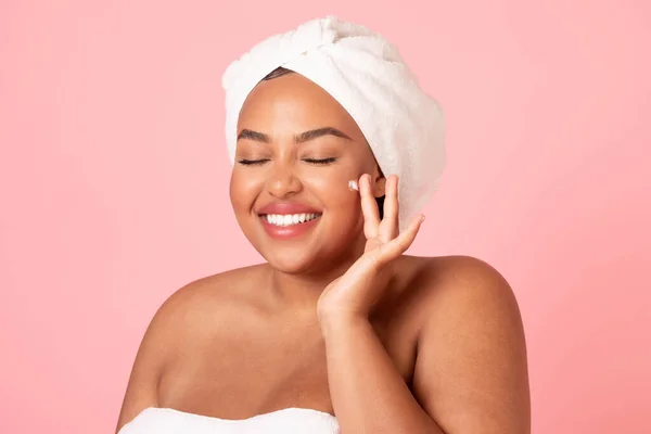 フェイスクリームを使用してタオルで包まれた女性は 彼女の顔と笑顔に美容製品を適用する幸せな黒体の正の女性は ピンクのスタジオの背景に隔離されました — ストック写真