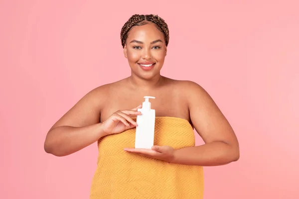 美しい黒肥満の女性は 保湿ボディローションとタオル保持瓶に包まれ 女性はピンクのスタジオの背景にポーズを 新しいスキンケア製品を使用して モックアップ — ストック写真