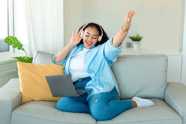 能量的情绪 快乐的黑人女性 戴着无线耳机 在笔记本电脑上听音乐 坐在舒适的沙发上 举手唱歌 — 图库照片
