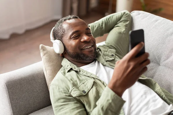 ワイヤレスヘッドフォンで幸せな中年アフリカ系アメリカ人男性は スマートフォンを入力し リビングルームのインテリアで余暇をお楽しみください オーディオアプリで休息とリラクゼーション 自宅でビデオを見る — ストック写真