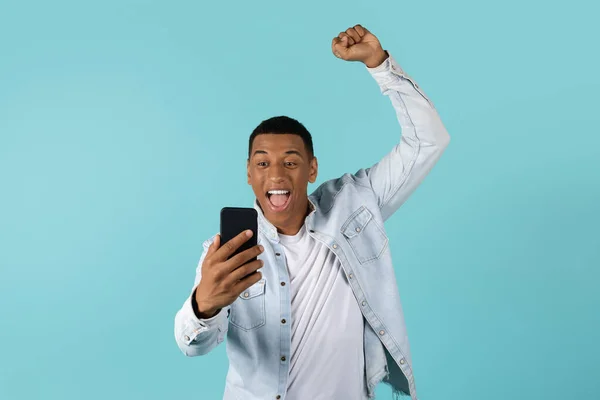 兴奋兴奋的年轻非洲裔美国男性 漫不经心地张开嘴看智能手机 举手表决胜利和成功的姿态 庆祝胜利 孤立在蓝色背景下 工作室 好消息 — 图库照片