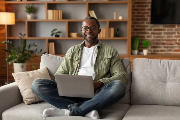 アフリカ系アメリカ人の中年男性の笑顔は ノートパソコンを入力し リモートで仕事をし ビデオを見て リビングルームのインテリアでソファの上に座っています 自宅でインターネットでのフリーランス ビジネス サーフィンのためのデバイス — ストック写真