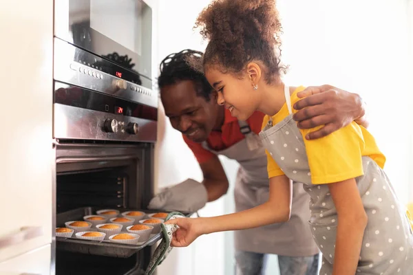 家庭で一緒にキッチンでマフィンを焼く幸せな黒父と十代の娘 笑顔アフリカ系アメリカ人の父と女性の子供はオーブンから焼きたてのペストリーでトレイを取ります クローズアップショット — ストック写真