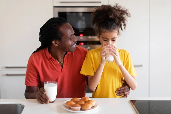 黒人のPreteen Girlと彼女の父は一緒に台所で軽食を食べています アフリカ系アメリカ人のお父さんと彼の小さな娘は自家製のマフィンを楽しんで ミルクを飲みます 自宅で一口を持っています フリースペース — ストック写真